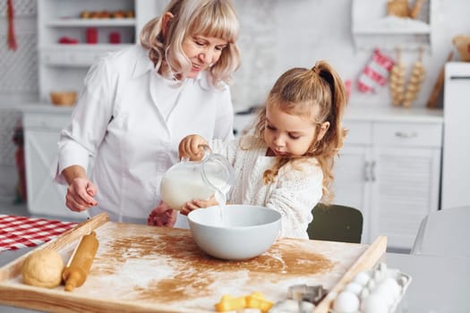 Fills with milk. Senior grandmother with her little granddaughter cooks sweets for Christmas on the kitchen.
