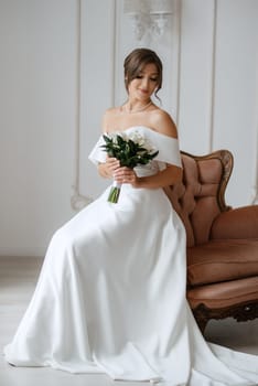 brunette bride in a tight wedding dress in a bright studio with a bouquet on an armchair
