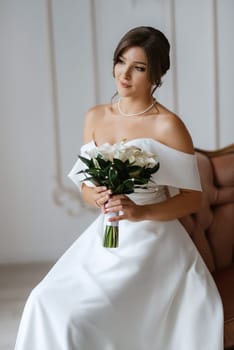 brunette bride in a tight wedding dress in a bright studio with a bouquet on an armchair