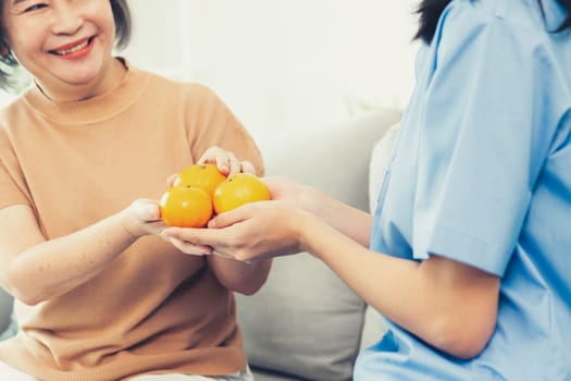 A young caregiver handing oranges to her contented senior patient at the living room. Senior care services, home visit by medical.