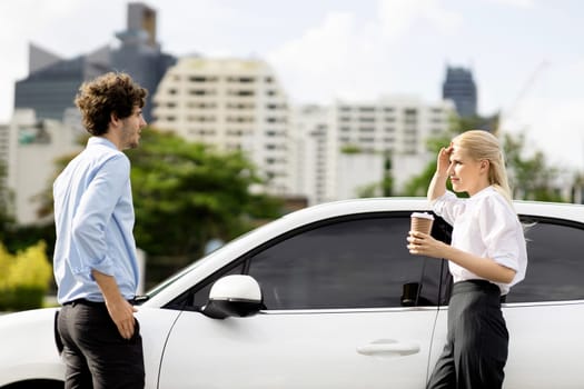 Progressive businessman and businesswoman with coffee, standing at electric car connected to charging station before driving around city center. Eco friendly rechargeable car powered by clean energy.