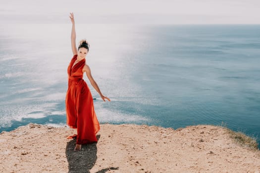 Side view a Young beautiful sensual woman in a red long dress posing on a rock high above the sea during sunrise. Girl on the nature on blue sky background. Fashion photo.