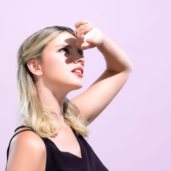 Closeup young blond hair woman with perfect skin and soft alluring facial makeup raise her hand cover her face from bright sunlight in pink isolated background for skincare sunscreen product.