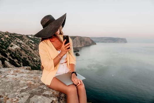 Successful business woman in yellow hat working on laptop by the sea. Pretty lady typing on computer at summer day outdoors. Freelance, travel and holidays concept.