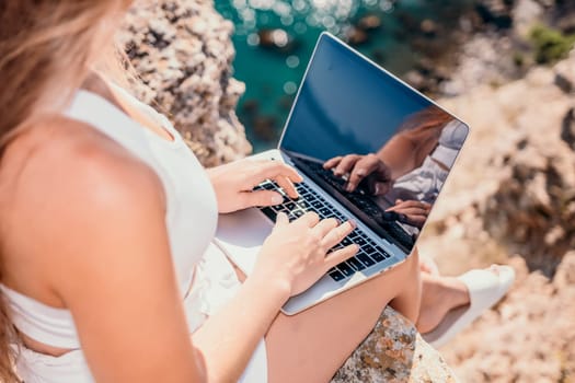 Successful business woman in yellow hat working on laptop by the sea. Pretty lady typing on computer at summer day outdoors. Freelance, travel and holidays concept.