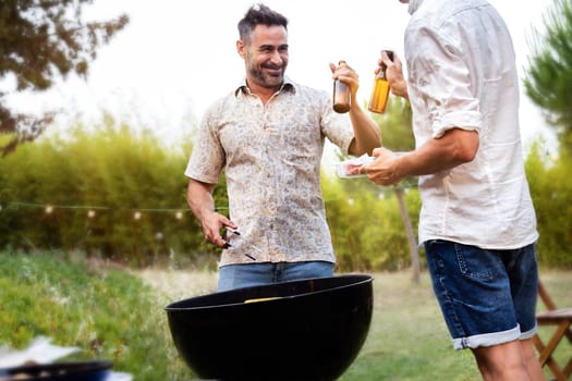 Two male friends toasting with beer while cooking food on grill. Outdoor garden barbecue party. Weekend activities concept.