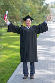 An elderly gray-haired man in a graduate robe spread his arms to the sides and holds a diploma outdoors. Vertical