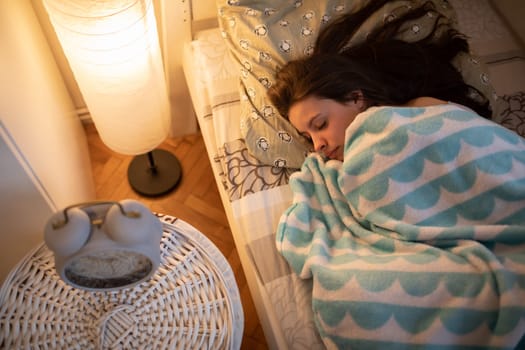 A young woman sleeps hunched over by the light of a bedside lamp. The light of a lamp on the wall. A girl under a soft blanket.