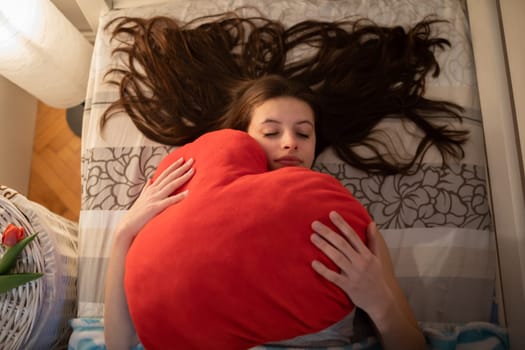 A red pillow in the hands of a sleeping girl. The light of a lamp on the wall.