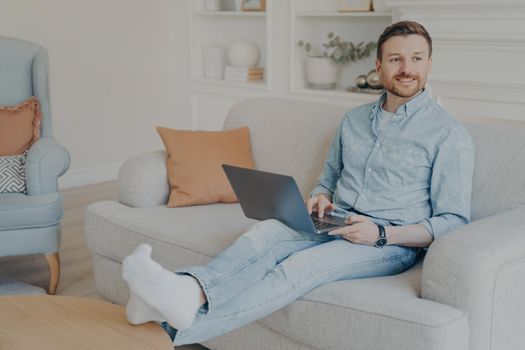 Young man with stubble surfing web on his laptop while sitting on couch, resting legs on coffee table, relaxing after hard work session on project, taking break from working