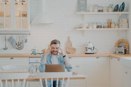 Handsome young businessman in casual clothes working with computer remotely while sitting at wooden table in stylish interior kitchen and smiling, looking at laptop screen with satisfied expression