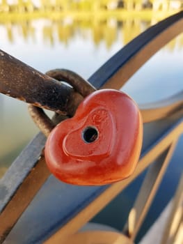 Red lock in the shape of a heart on the metal fence of the city embankment. The lock is metal hinged.