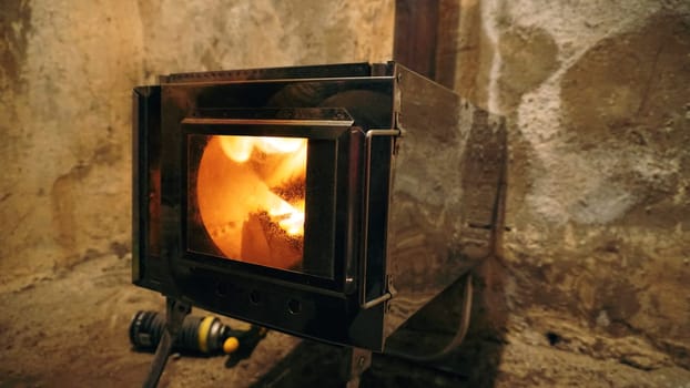 A fire is burning in a camping stove with a glass door. The process of smoldering fuel briquettes is visible. The lining of the stove made of mirror metal reflects the gray old walls of the hut.