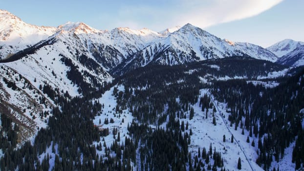 Coniferous forest in snowy mountains. White clouds float across the sky. Top view from a drone. In places, the road to the mountains and buildings are visible. There is a pipe along the gorge. Sunset