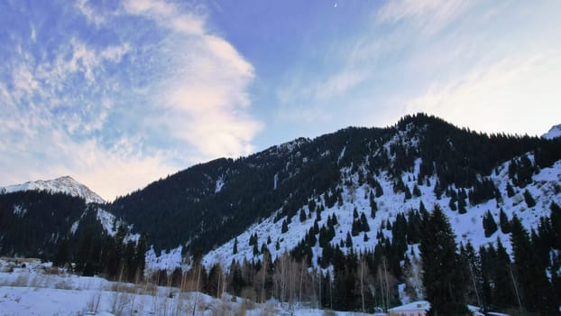 Coniferous forest in snowy mountains. White clouds float across the sky. Top view from a drone. In places, the road to the mountains and buildings are visible. There is a pipe along the gorge. Sunset