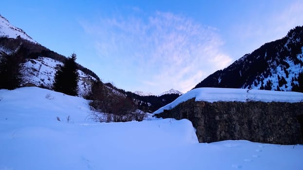 An old stone prison or hut in the mountains. Snowy mountains, forest and clouds against a blue sky. Tall dry bushes peek out from under the snow. Birch and spruce trees are visible in the distance.