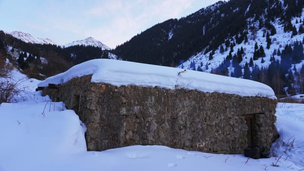 An old stone prison or hut in the mountains. Snowy mountains, forest and clouds against a blue sky. Tall dry bushes peek out from under the snow. Birch and spruce trees are visible in the distance.