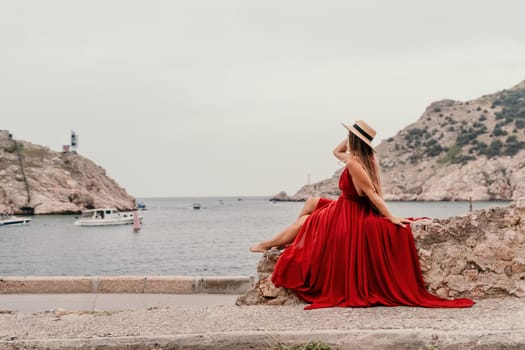 Side view a Young beautiful sensual woman in a red long dress posing on a volcanic rock high above the sea during sunset. Girl on the nature on overcast sky background. Fashion photo