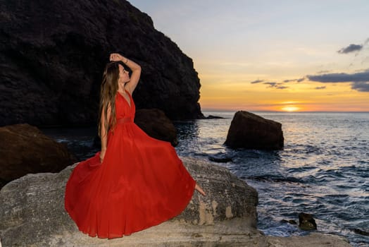 Woman in a red flying dress on the ocean or on the sea beach against the backdrop of the sunset sky