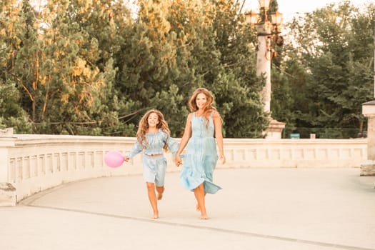 Daughter mother run holding hands. In blue dresses with flowing long hair against the backdrop of a sunset and a white building