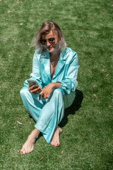 a young girl with blond curly hair in glasses and in a blue overalls lies on the summer fresh green grass with a phone in her hands. open image
