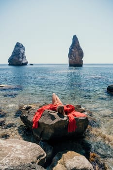 Woman travel sea. Young Happy woman in a long red dress posing on a beach near the sea on background of volcanic rocks, like in Iceland, sharing travel adventure journey