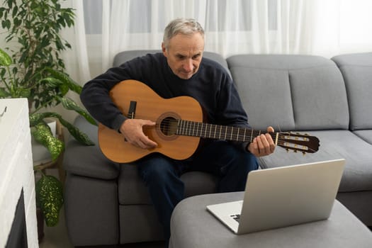 Portrait of senior man in headphones taking online guitar lesson looking at laptop screen. Retired male learning to play guitar watching webinar on computer at home.