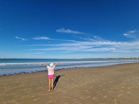 Girl with hat opening her arms in front of the sea with a seagull flying in front of her, on a sandy beach. Valdelagrana beach in El Puerto de Santa Maria, Cadiz, Andalusia, Spain.