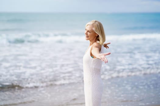 Mature woman opening her arms on a tropical beach, spending her leisure time. Elderly female enjoying her retirement at a seaside retreat.