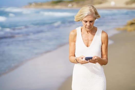 Elderly female in summer dress standing on sandy seashore and messaging on mobile phone on sunny day