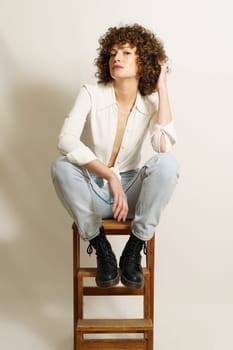 Full length of flirty woman model with curly hair, in stylish wear resting on wooden stool playing with curl and looking at camera against white background