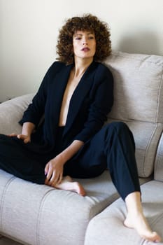 Full body high angle of adult woman with curly dark hair sitting on couch wearing black jacket and pants looking at camera