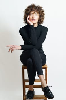 Concentrated and confident young female, in black outfit with hand touching cheek sitting on chair with legs crossed while leaning on elbow and looking at camera in gray backdrop