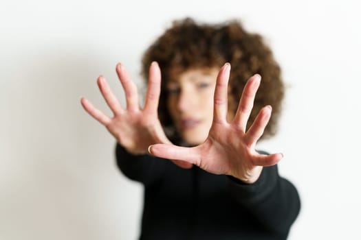 Defocused face of girl model, with curly hair looking at camera and showing stop gesture with full open palms and fingers against gray background in blurring unseen side illuminating lights