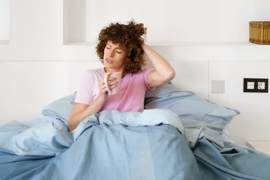 Lady touching head with closed eyes while sitting on bed and drinking hot beverage in morning