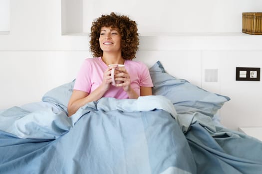 Positive female with curly hair sitting on bed and looking away while drinking tea in early morning