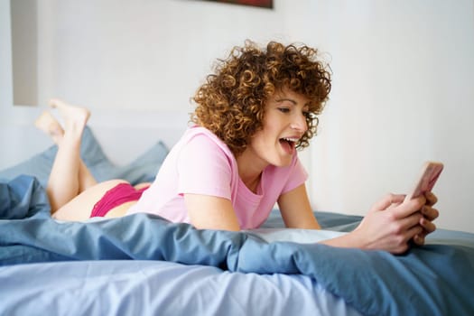 Full body side view of cheerful young female with curly dark hair lying on bed and browsing mobile phone during weekend at home