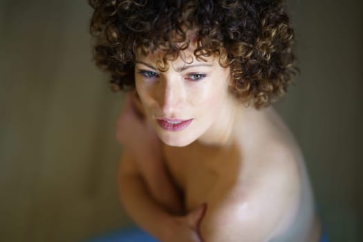 High angle of pensive young nude female with curly hair looking away while sitting on floor with crossed arms