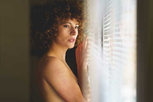 Side view of charming young naked curly haired female leaning on window and looking away thoughtfully in light room