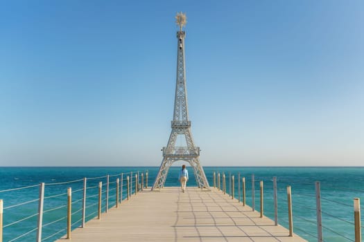 Large model of the Eiffel Tower on the beach. A woman walks along the pier towards the tower, wearing a blue jacket and white jeans