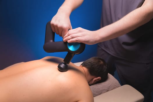 Therapist massaging a man's back with a percussion massage device in a massage room. The therapist's hand holds a therapeutic vibratory massager. Physiotherapy and Muscle Recovery and Massage.