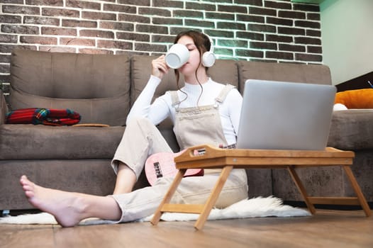 Pretty young woman with a laptop sits on the floor at home. Side view, young female freelancer in home overalls sits on the floor and uses a laptop computer with a cup of hot drink at home. Work from home concept.