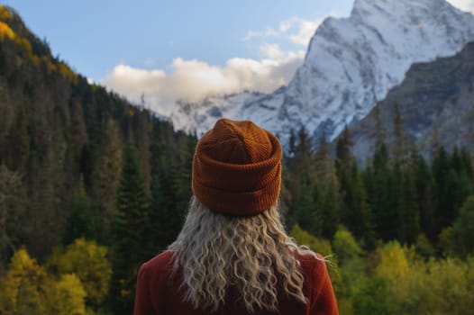 Blonde woman standing with her back to the camera and watching magical clouds in a mountainous area. Woman in a hat in summer or autumn in the snowy mountains next to the green forest.