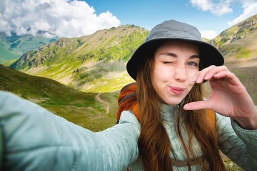 Cute happy girl making heart shaped hand gesture looking at camera smiling funny, caucasian young single woman blogger laughing face showing love sign symbol, mountains, nature, closeup portrait.