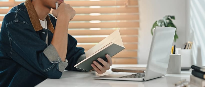 Stylish male hand holding notebook, working with laptop computer in home office. Cropped shot.