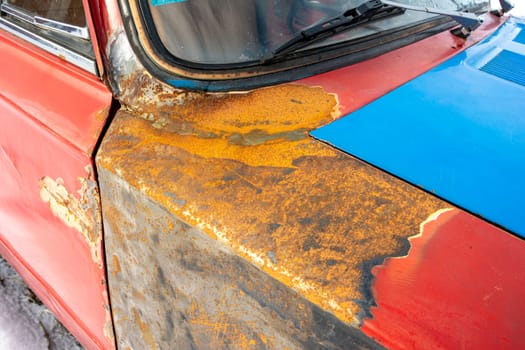 the wing of an old rusty car in close-up. the red door, rusty fender, windshield and blue hood are partially visible. restoration of vehicles.