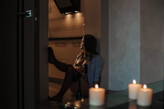 beautiful woman sits in the kitchen with candles in a man's shirt and drinks wine.