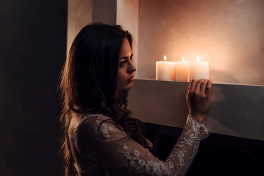 beautiful brunette in a white transparent tunic stands near the candles.