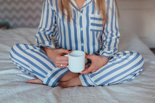 In the morning, a girl in pajamas sits on the bed with a cup of coffee in her hands . coffee in bed concept