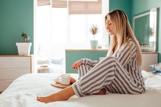 a woman in pajamas drinks coffee with a croissant on the bed . the concept of breakfast in bed.